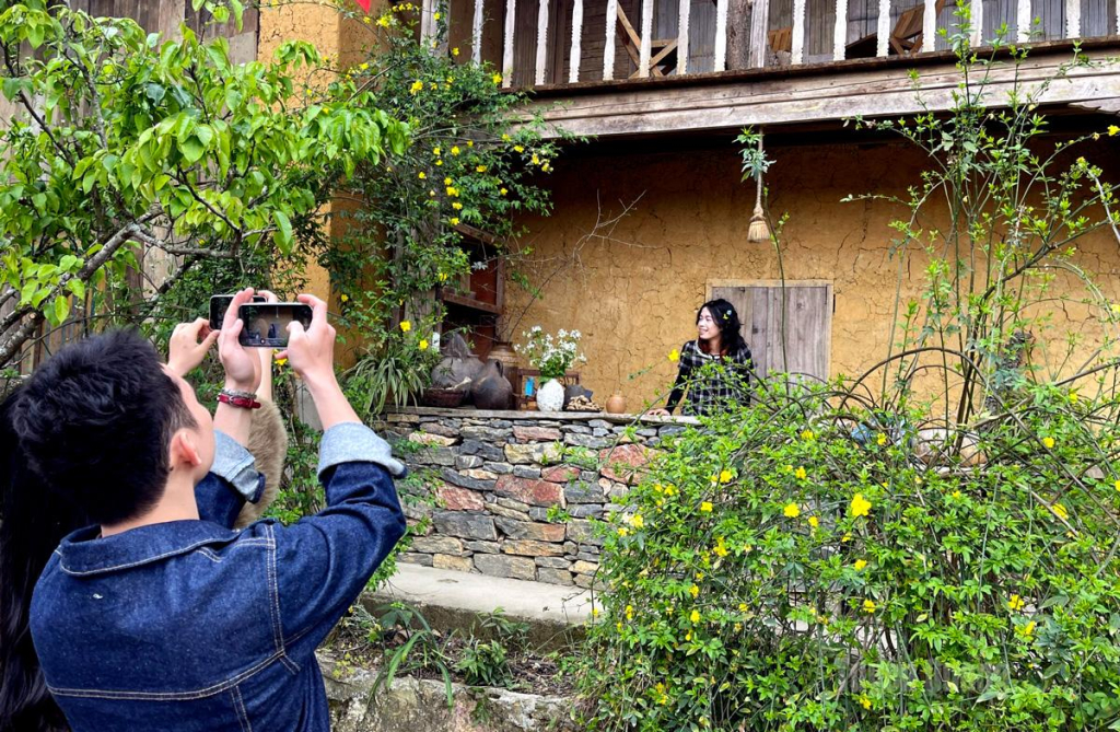 Visitors pose beside the porch of a traditional rammed-earth house in Lo Lo Chai Village, where bright yellow flower trellises and the rustic setting create an attractive check-in spot.